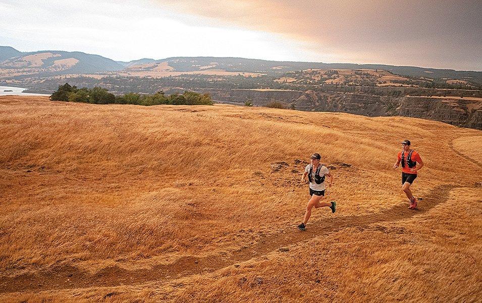 A woman in a white shirt and trail running shorts jogs on a scenic path ahead of a man wearing a red shirt.