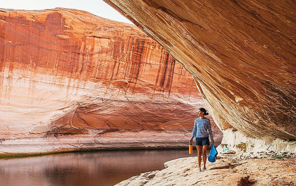 A woman in a blue striped shirt hikes through a stunning slot canyon with a yellow water bottle in one hand and a blue dry bag in the other. 