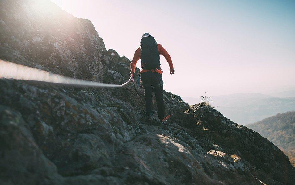 Using a via ferrata cable for stability, a trail runner begins ascending a steep, rocky incline. 