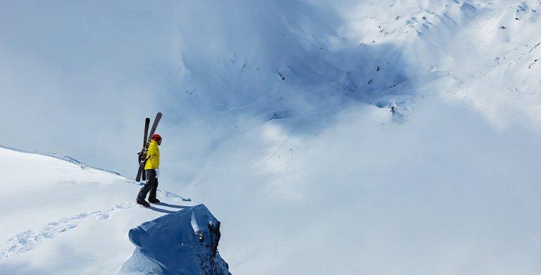  A person has boot-packed to the edge of a snow-covered cliff, skis attached to his backpack. He pauses, pondering whether to ski down or retrace his steps?