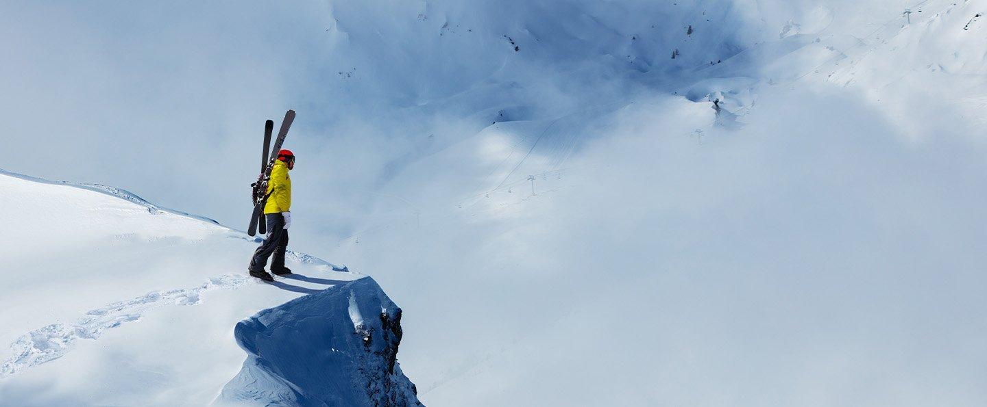  A person has boot-packed to the edge of a snow-covered cliff, skis attached to his backpack. He pauses, pondering whether to ski down or retrace his steps?