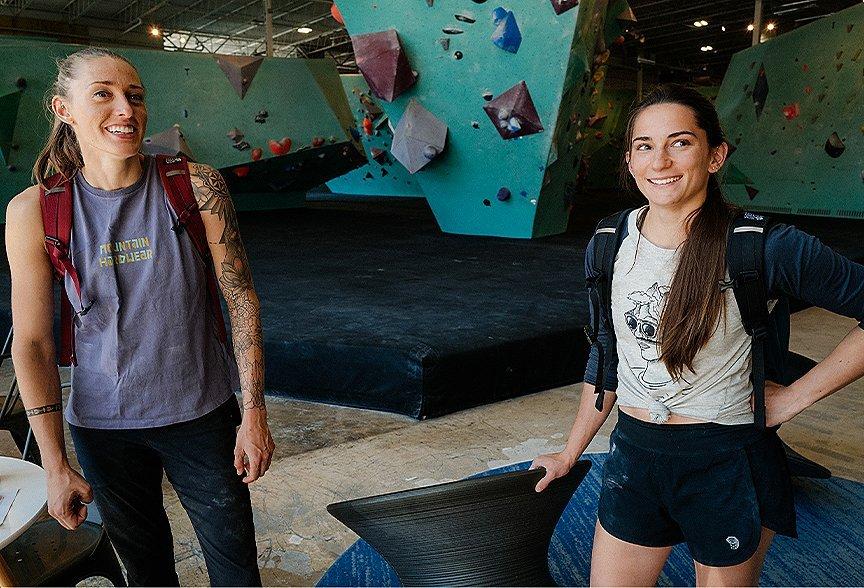 Alex Johnson and Kyra Condie smile for the camera inside the climbing gym, both with backpacks on, ready to go.