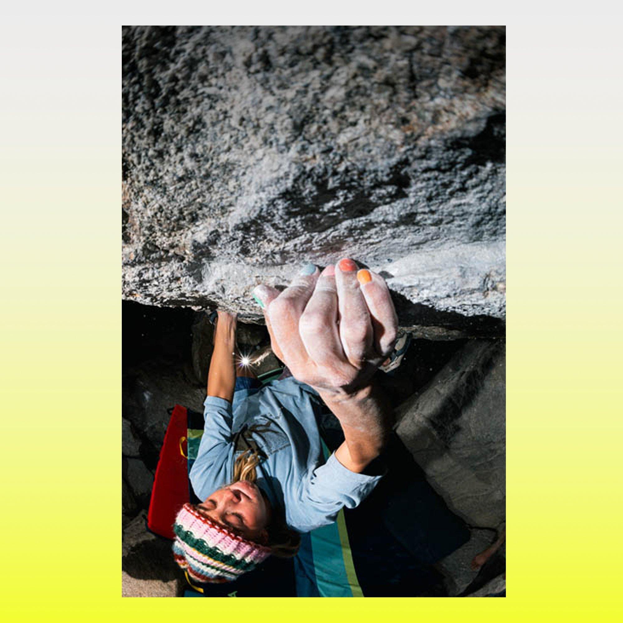 A woman with chalky hands grimaces with focus while bouldering in a Mountain Hardwear tee and colorful knit beanie above a crash pad.