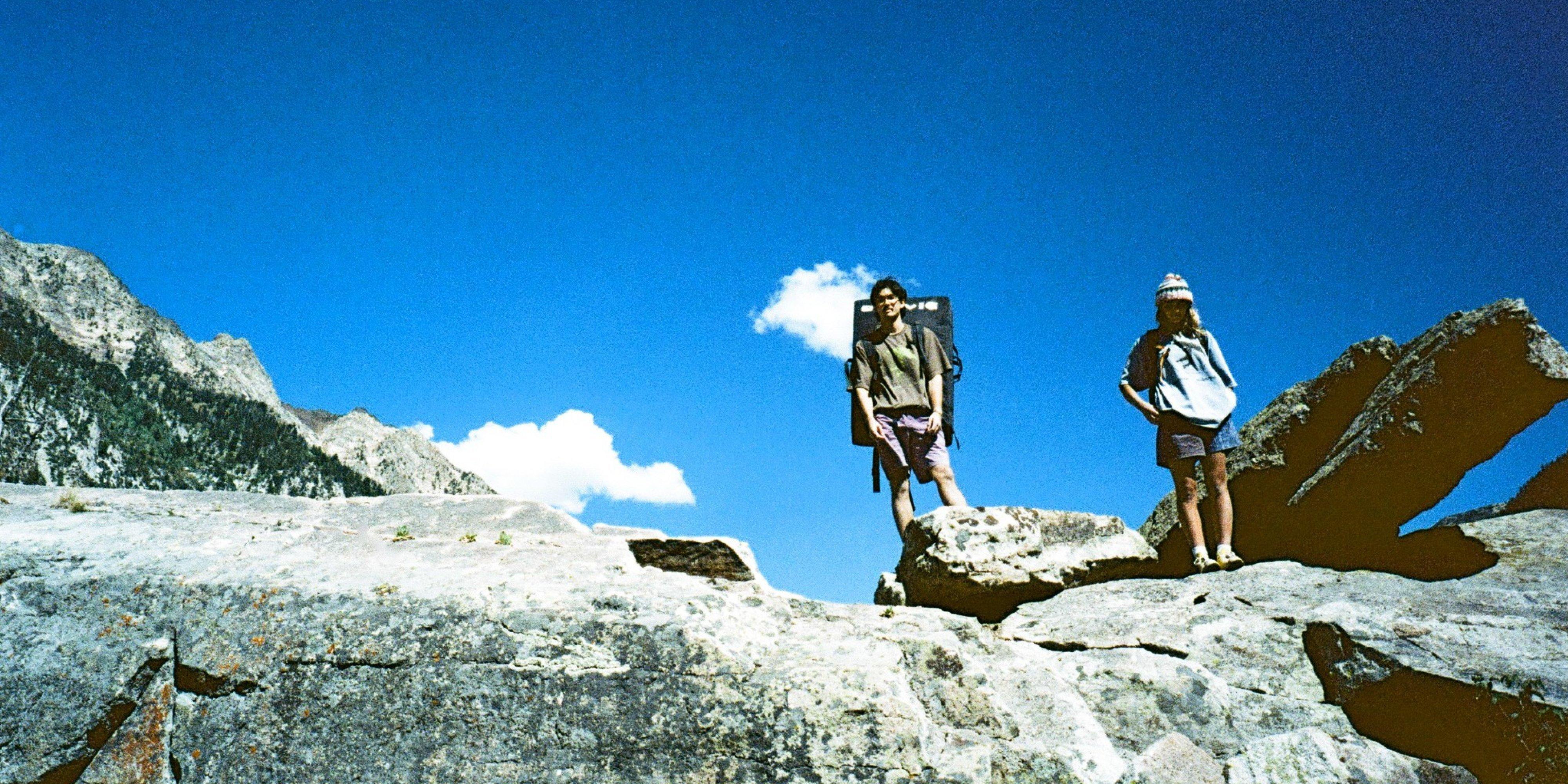 Two climbers in Rockrydge™ Shorts and graphic tees, one with a crash pad on his back, peer down from atop a jagged boulder. 