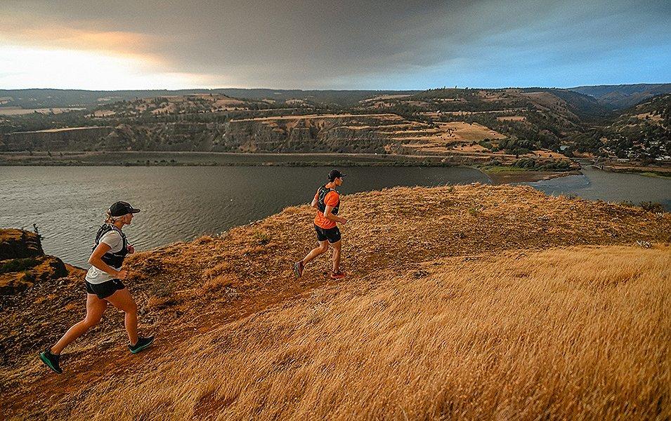 A man wearing an orange Columbia Sportswear shirt and vest runs along a scenic trail with a woman in a white shirt jogging behind him.