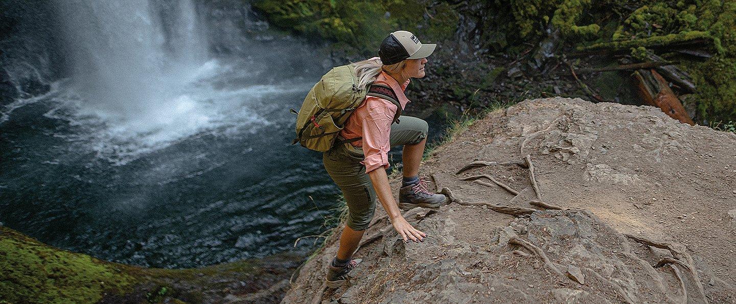 A woman wearing a pink hiking shirt and pale green pants climbs up a rock in a scenic forest with a waterfall in the background. 