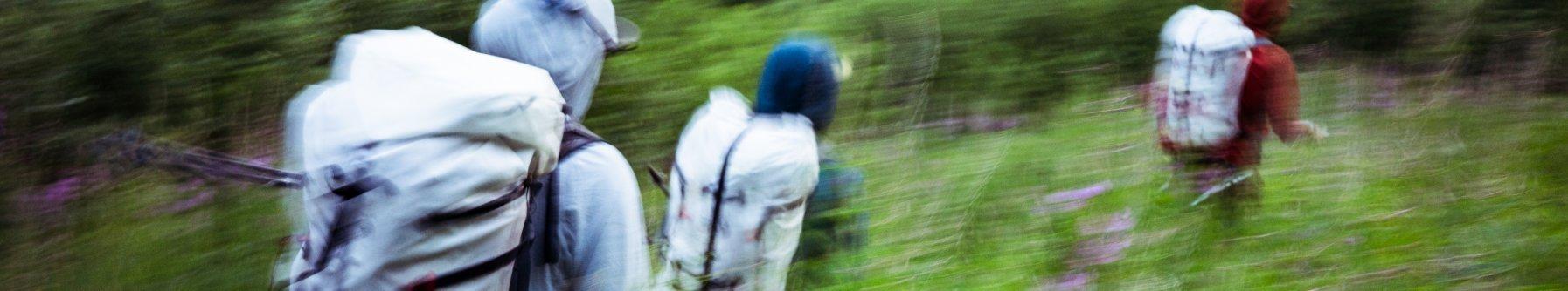 Abstract motion blur image of hikers crossing a stream, wearing large backpacks and outdoor gear. The photo captures dynamic movement with streaks of color, emphasizing speed, activity, and the energy of backcountry adventure.