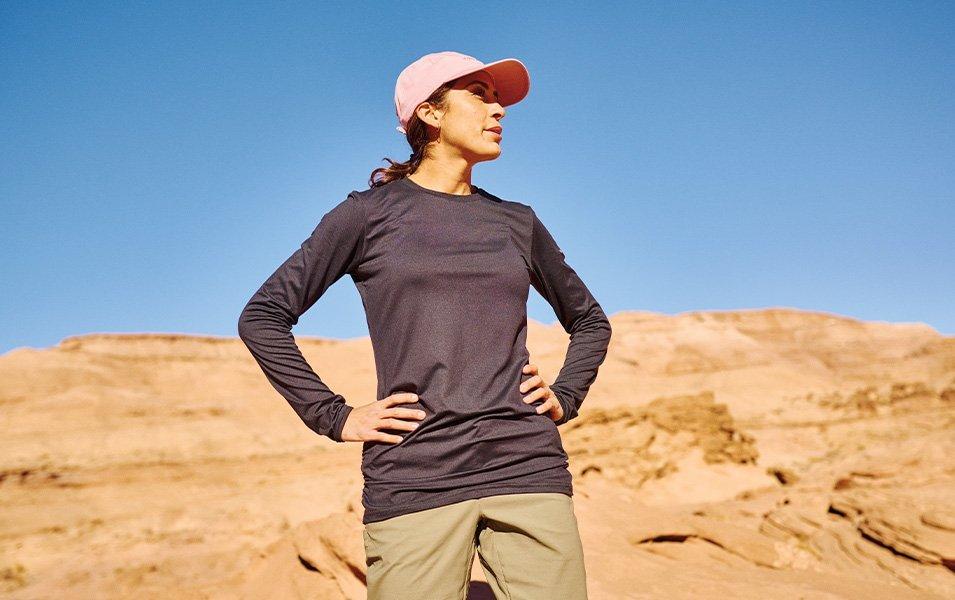 A woman wearing a brimmed ball cap and a long-sleeve sun-protective shirt stands with her hands on her hips. 