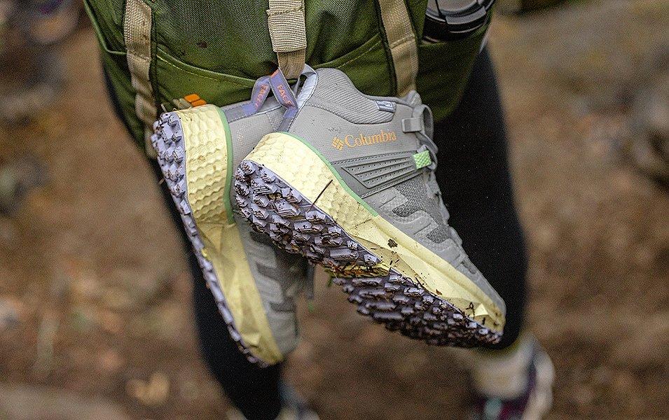A pair of Columbia Sportswear hiking boots hang from a hiker’s backpack as they walk on a trail 