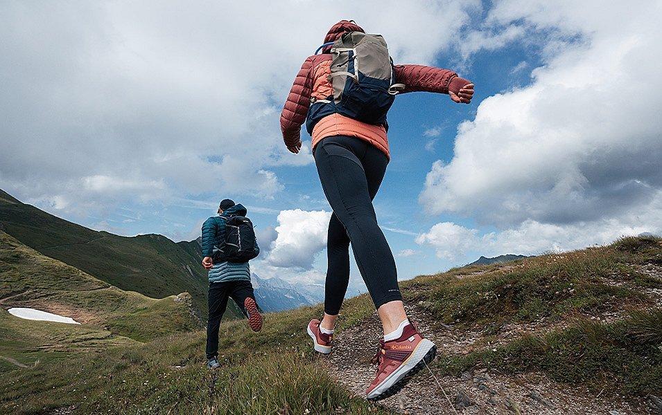 Two hikers in puffy jackets and small backpacks hike swiftly along a ridge-top trail.