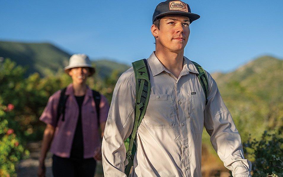 Two hikers stroll along a trail with rolling hills in the background and bushes with flowers in the foreground.