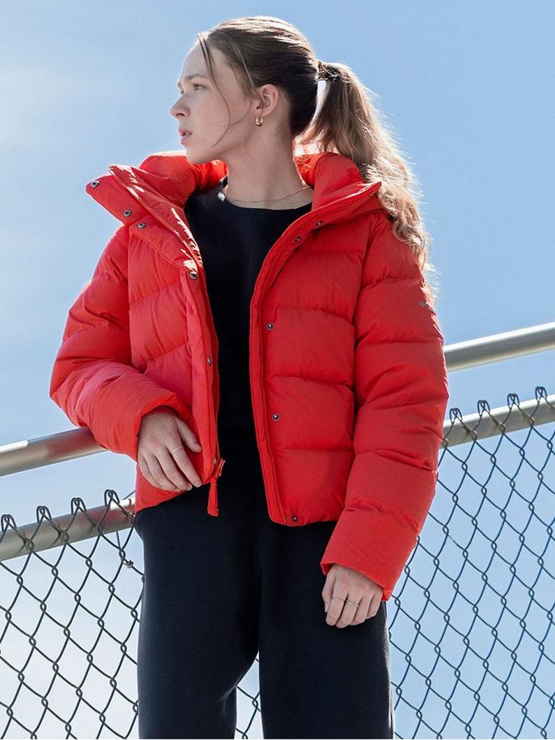 Woman wearing red puffer jacket with hood over black outfit, standing outdoors near chain-link fence.