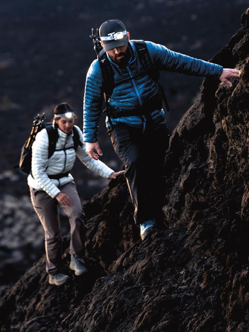 Two hikers wearing headlamps and insulated jackets climbing rocky terrain with backpacks and trekking poles.