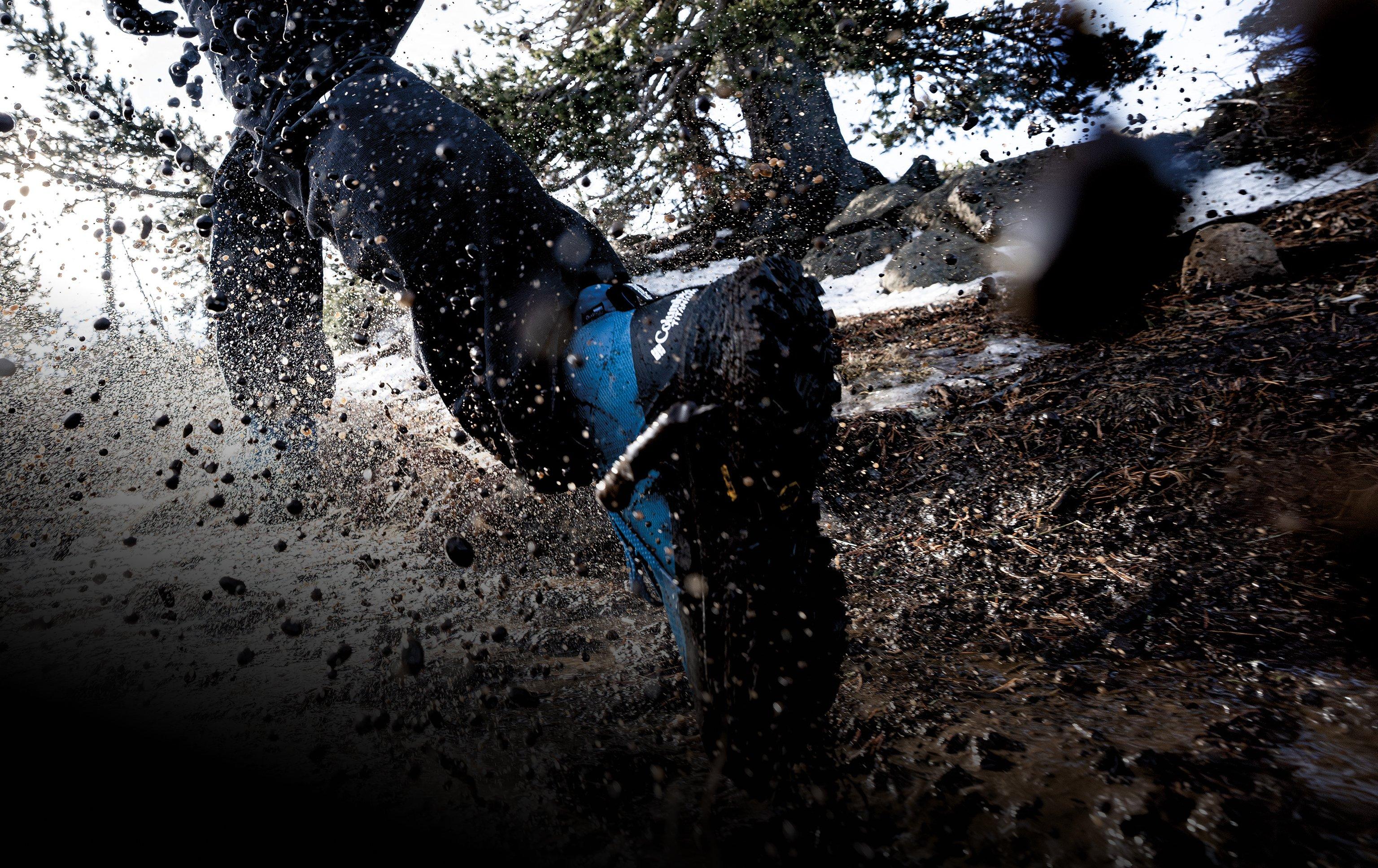 Person running through muddy water wearing blue and black trail running shoes, splashing mud and water.