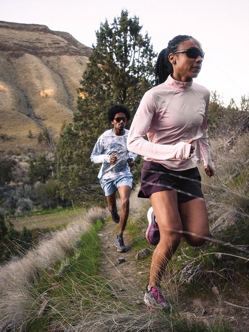 Woman in pink quarter-zip and black skort running on mountain trail, followed by man in tie-dye shirt.
