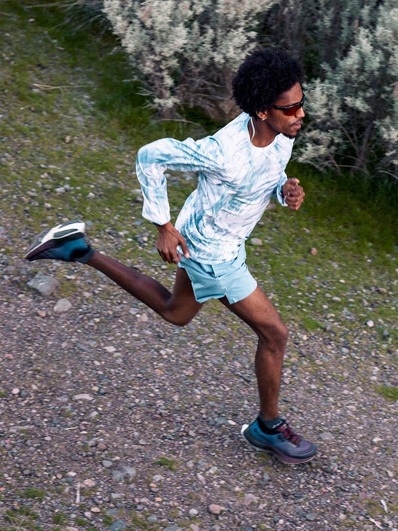 Man running on a gravel trail wearing blue tie-dye long sleeve shirt, light blue shorts, and sneakers.