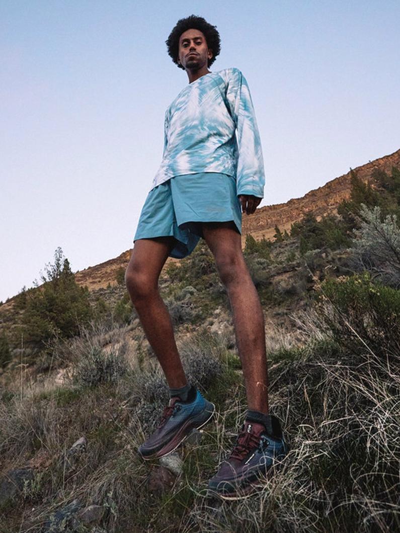 Man standing on rocky hillside wearing blue tie-dye sweatshirt, turquoise shorts, and hiking boots.