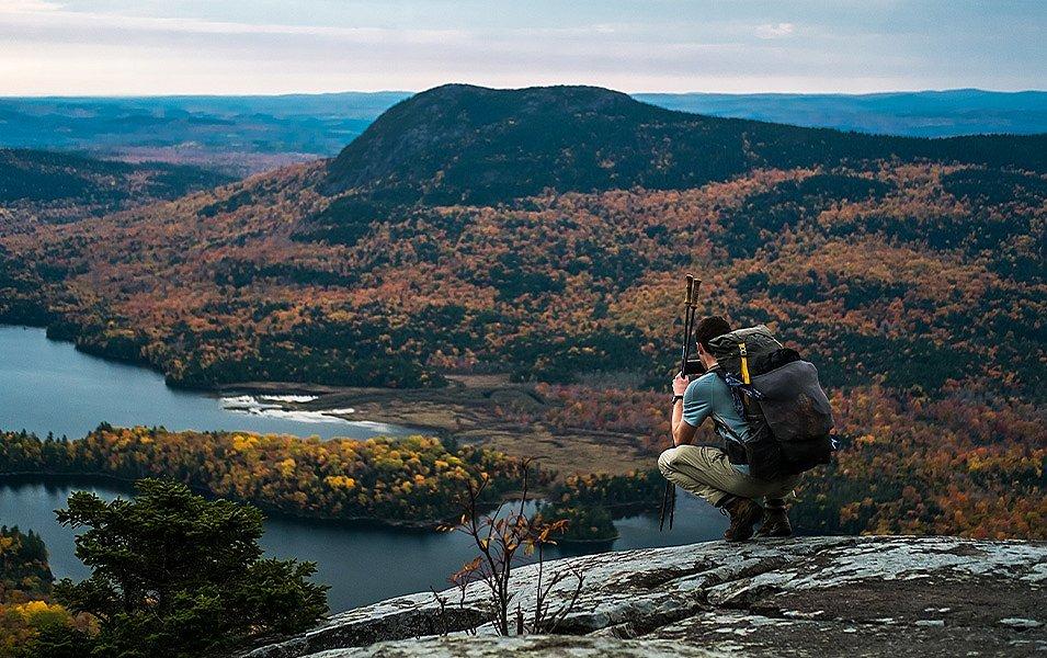 A hiker with a big backpack and hiking poles crouches on a rocky ridge, photographing a lake and tree-covered knoll.