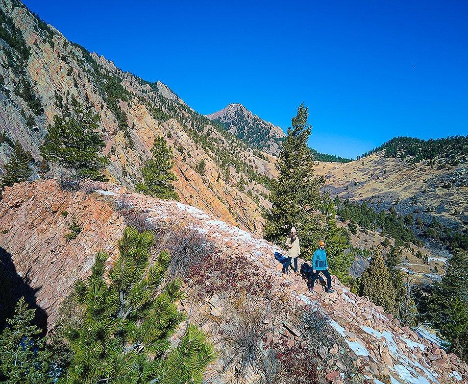 A pair of hikers walk down a steep, rocky trail in a beautiful mountainous setting. 