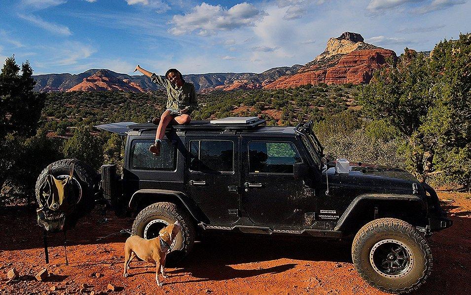 A woman on an overlanding adventure sits on top of a Jeep in the high desert smiling with her arm in the air as her dog looks up at her. 