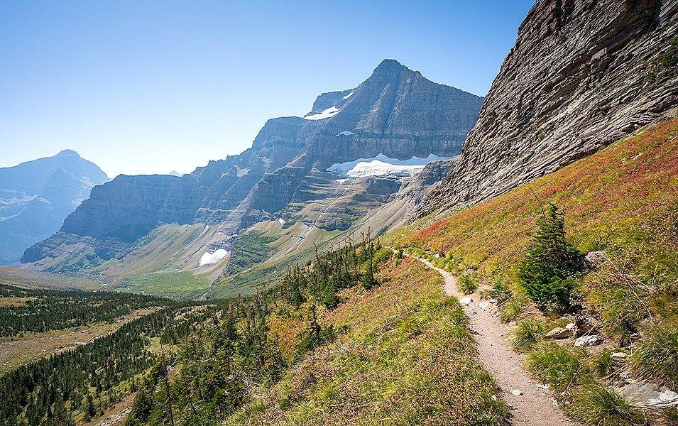 A dirt footpath traverses an alpine slope. The trail leads into a wide valley surrounded by steep mountains with several snowfields high up on their flanks.