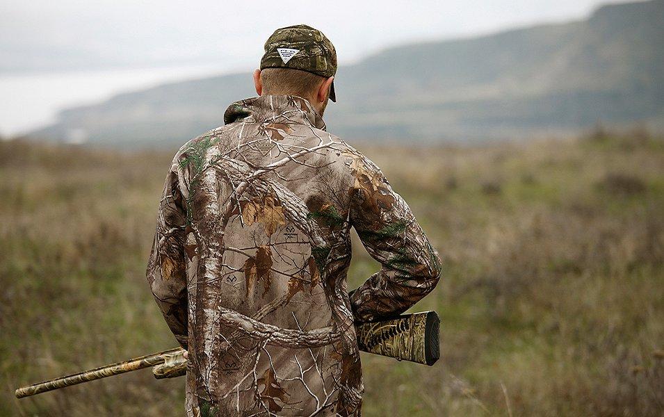 Un chasseur se tient dans un champ, tenant un fusil, dos à la caméra, portant des vêtements Columbia PHG.