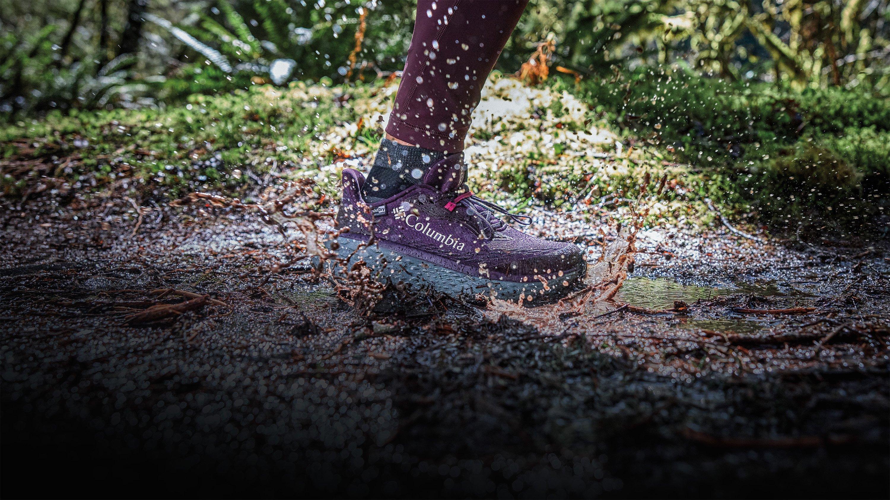 Purple hiking shoe splashing through muddy puddle on outdoor trail.