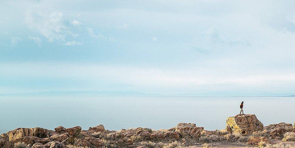 Man standing on a distant rock in front of a large lake.
