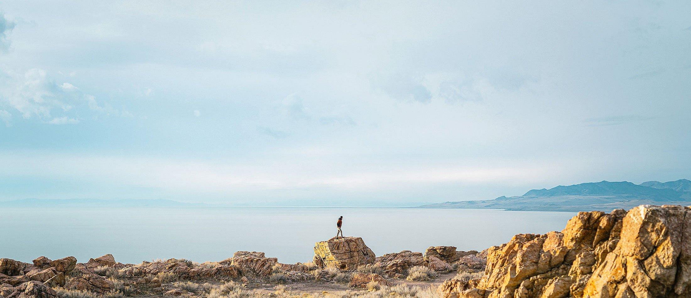 Man standing on a distant rock in front of a large lake.