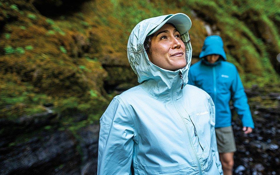 A woman in a light blue Columbia Sportswear rain jacket looks up in the middle of a wet, mossy forest as a man stands behind her looking down.
