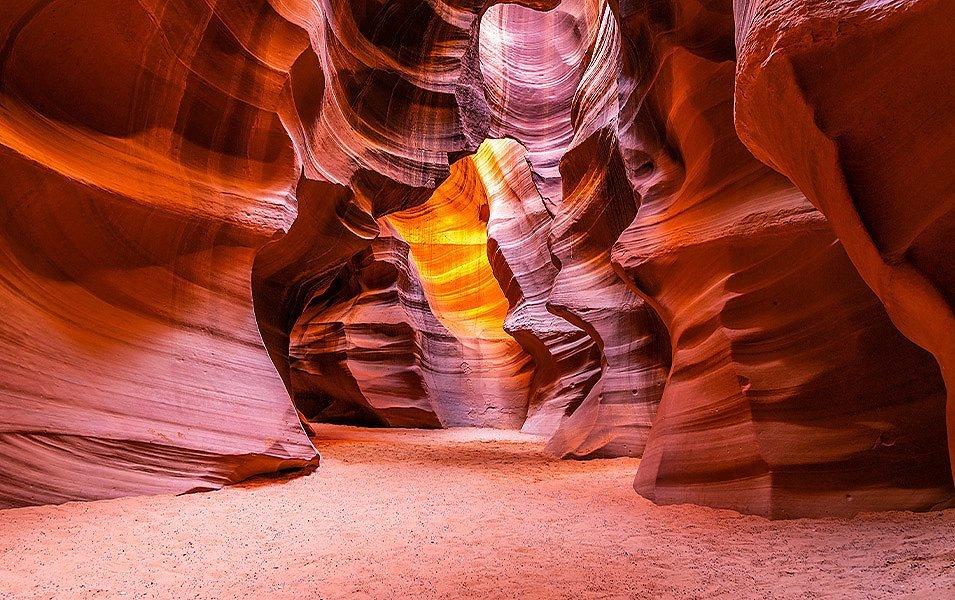 A narrow sandstone slot canyon with a sandy floor is illuminated from rays of light shining down from above. 