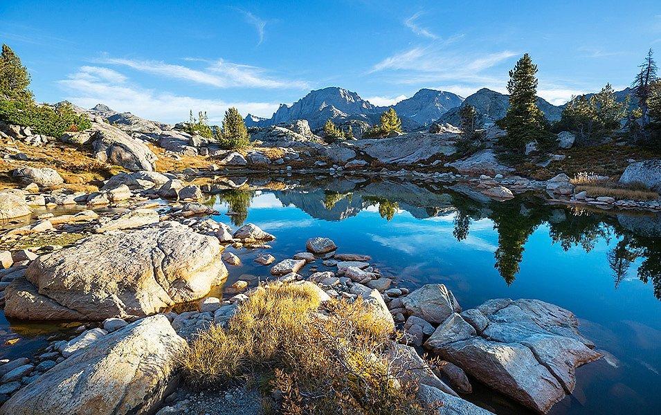 An azure lake is surrounded by scattered pine trees and granite boulders. A rocky mountain range is in the background.