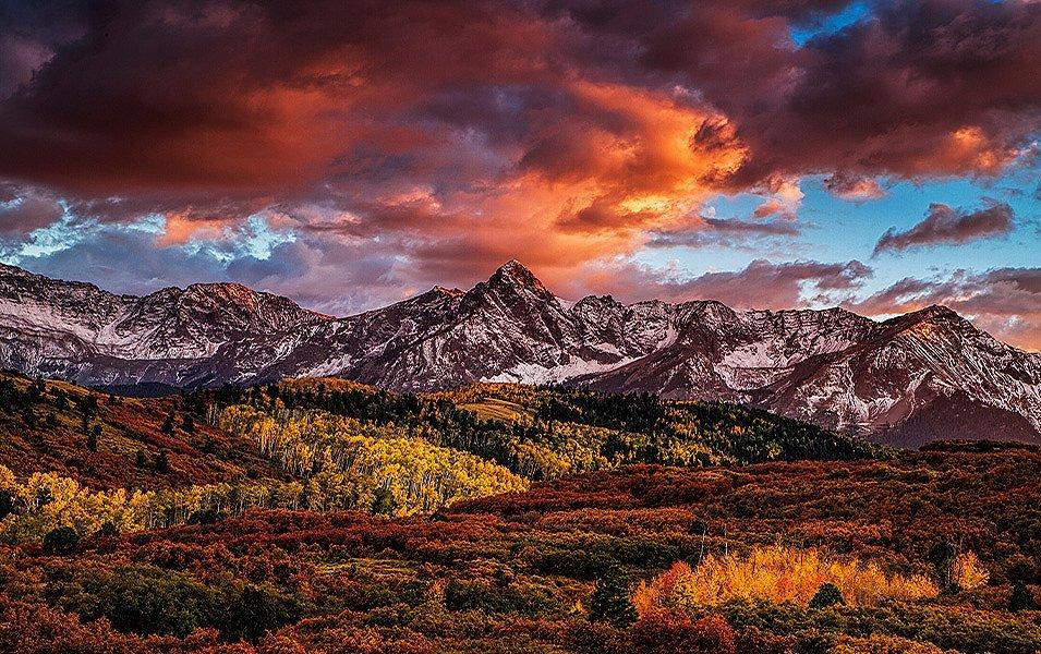 Winter is coming to this scenic valley. Stands of trees with orange and red leaves punctuate the foreground, and a mountain range looms in the background.