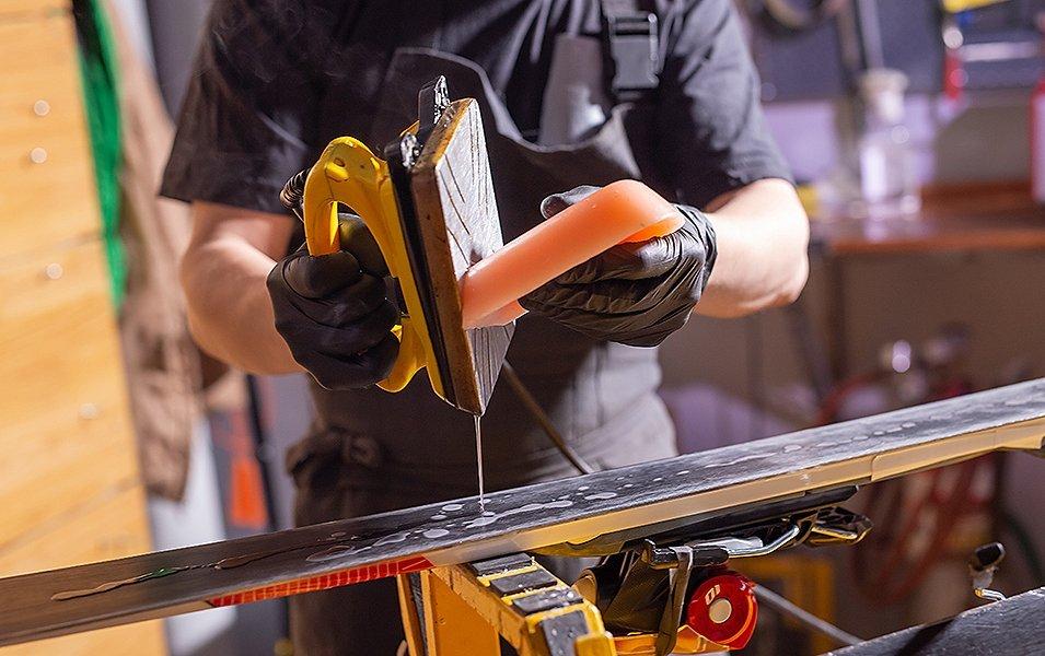 A man wearing black gloves holds a yellow iron as he drips wax onto a set of skis below.