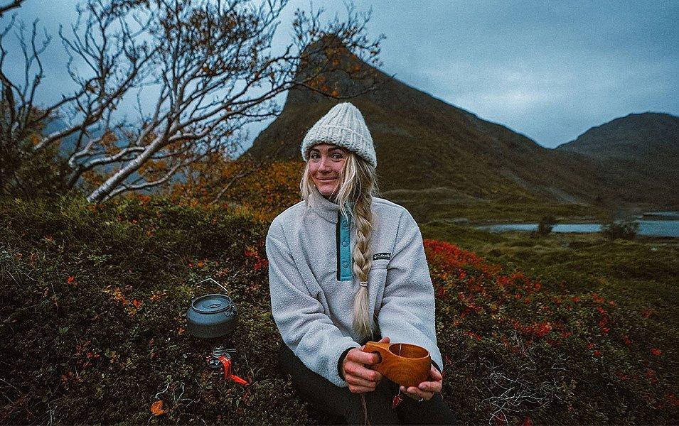  Rachel Pohl sits on a hillside in Norway wearing a white Columbia Sportswear fleece and holding a warm beverage as she smiles at the camera.