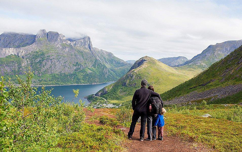 A friluftsliv-loving family stands on a scenic trail by the water staring at the beautiful fjords in the distance. 