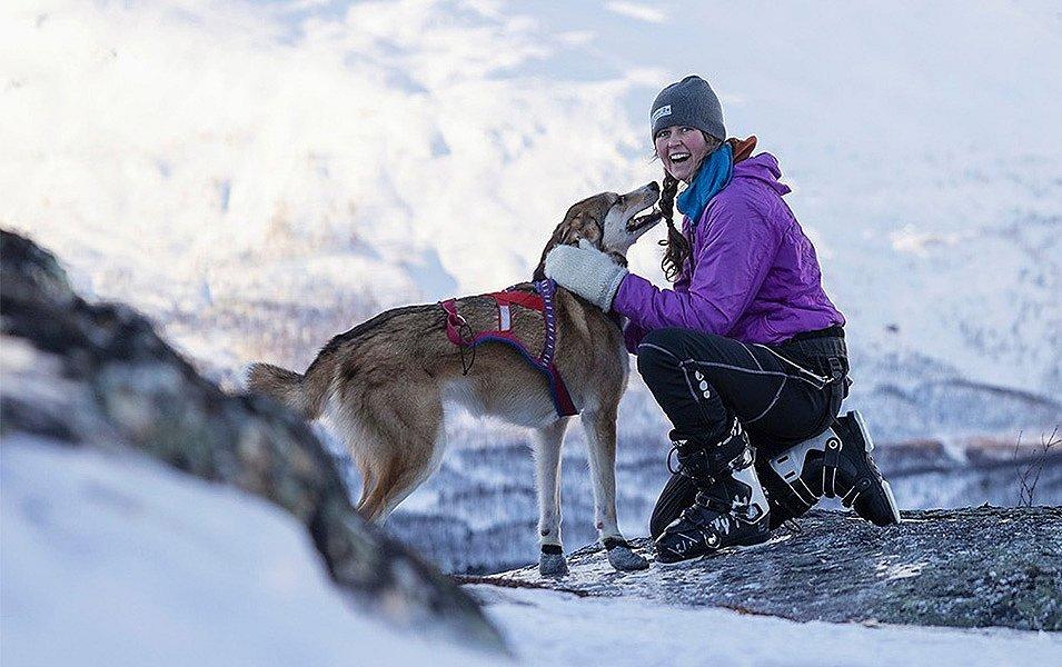  Magda Blakeson crouches in the snow petting a dog in front of a beautiful Norwegian backdrop. 