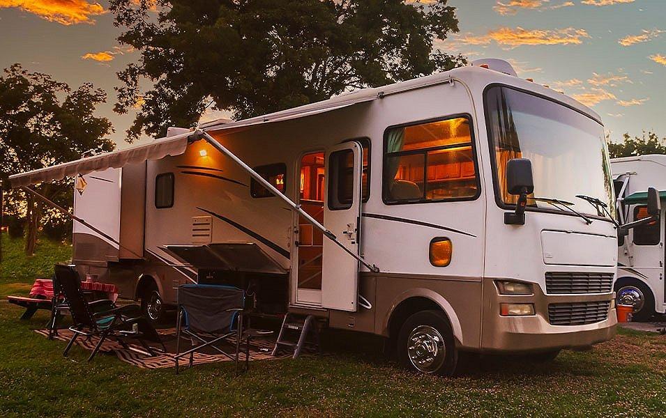 A photograph of a white RV sitting in a grassy area with an awning over some camping chairs. 