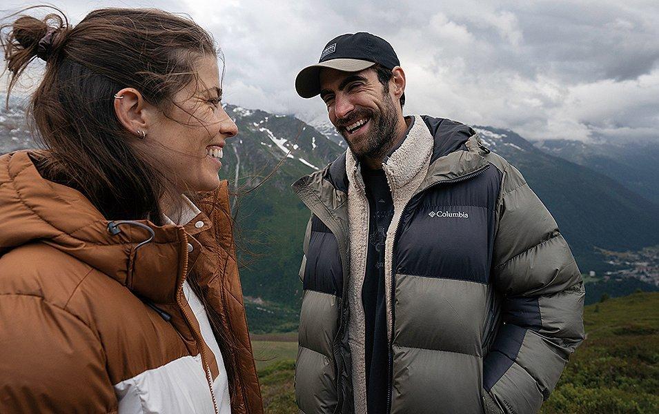 Smiling man and woman wearing Columbia puffer jackets on a grassy hilltop. 