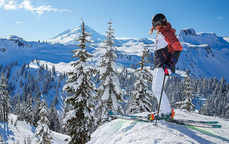 A skier wearing a red jacket and white ski pants stands on top of a steep mountain looking at the camera and smiling. 