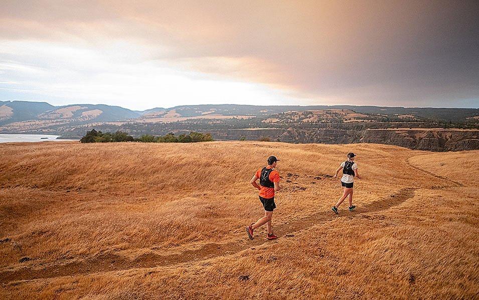  A man in a red shirt jogs behind a woman wearing white as they run along a narrow trail through an open field near a river. 