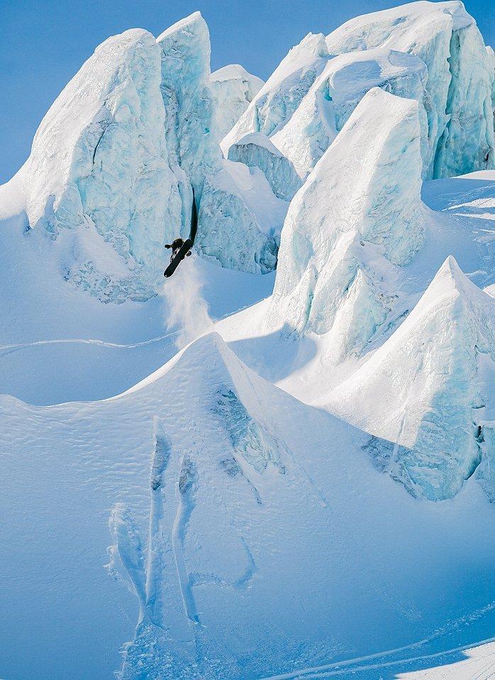 A snowboarder descends through steep, icy blue cliffs surrounded by deep snow, carving a dynamic path as powder flies up around them. The scene is framed by jagged, glacier-like formations under a clear blue sky, emphasizing the rugged and remote location