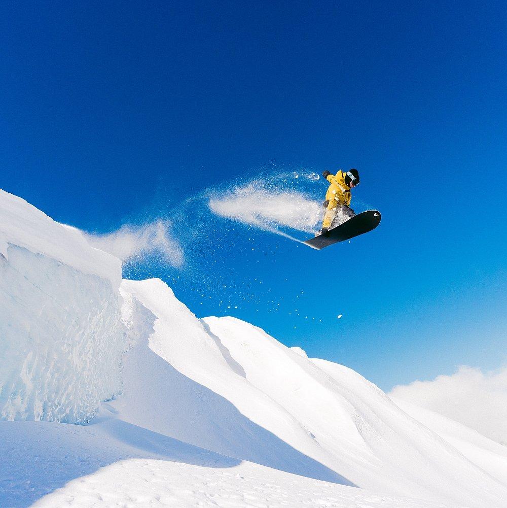 A snowboarder in a bright yellow suit catches air off a snow-covered ledge, leaving a trail of powder against a vibrant blue sky.