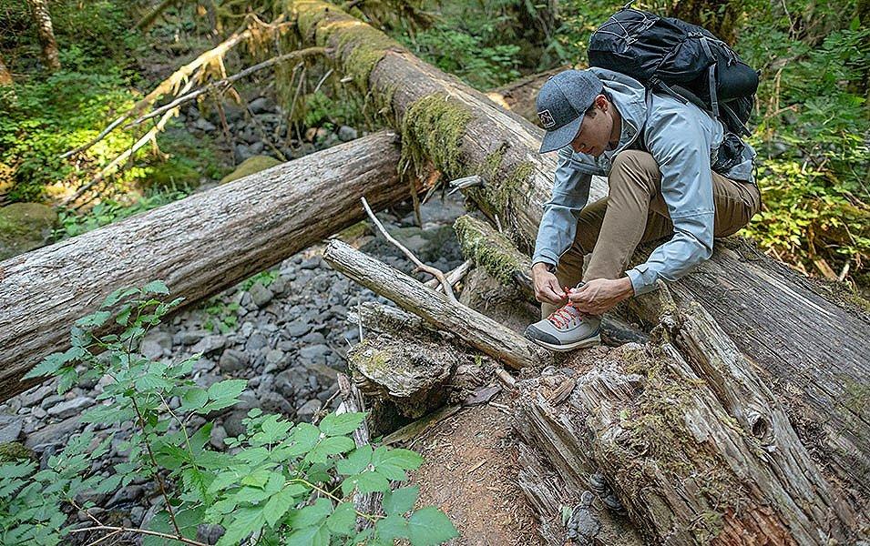 A man wearing a light blue Columbia Sportswear jacket stoops down on a mossy log in the woods to lace his hiking boots. 
