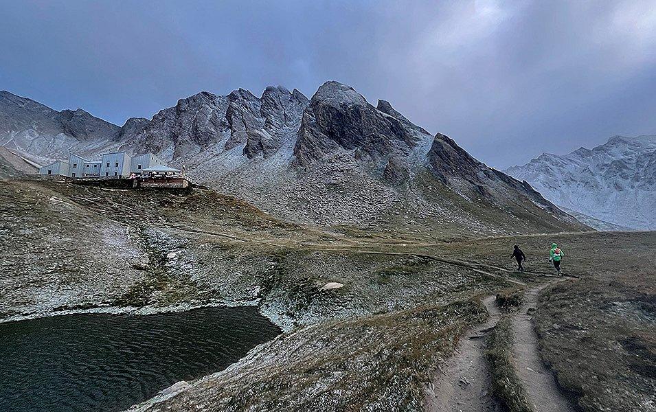 Two runners near a remote lodge built into the treeless alpine terrain above a lake. The runners are competing in the 200-mile Tor des Géants.  