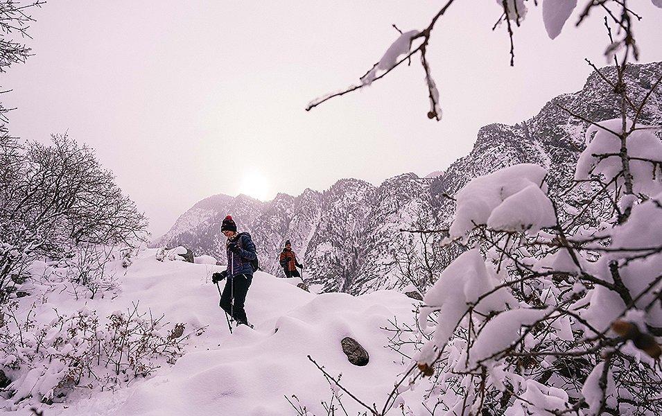 A woman treks through a snowy winter wonderland with another hiker and a stretch of mountains in the background. 