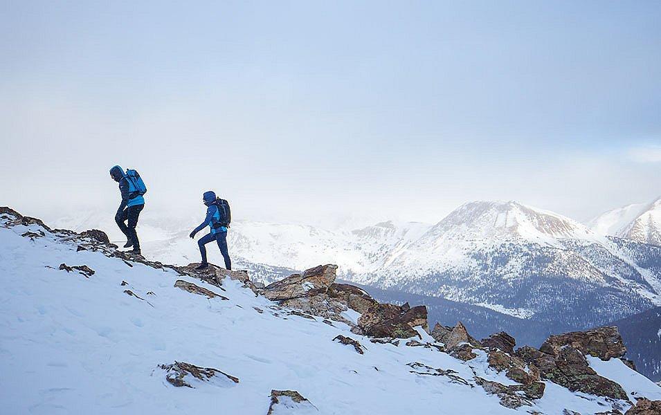 A pair of hikers wearing blue puffer jackets trek up a mountain trail in front of a scenic winter backdrop.  