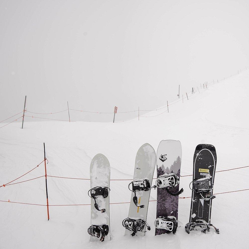 Snowboards lined up in the snow leaning against a fence.