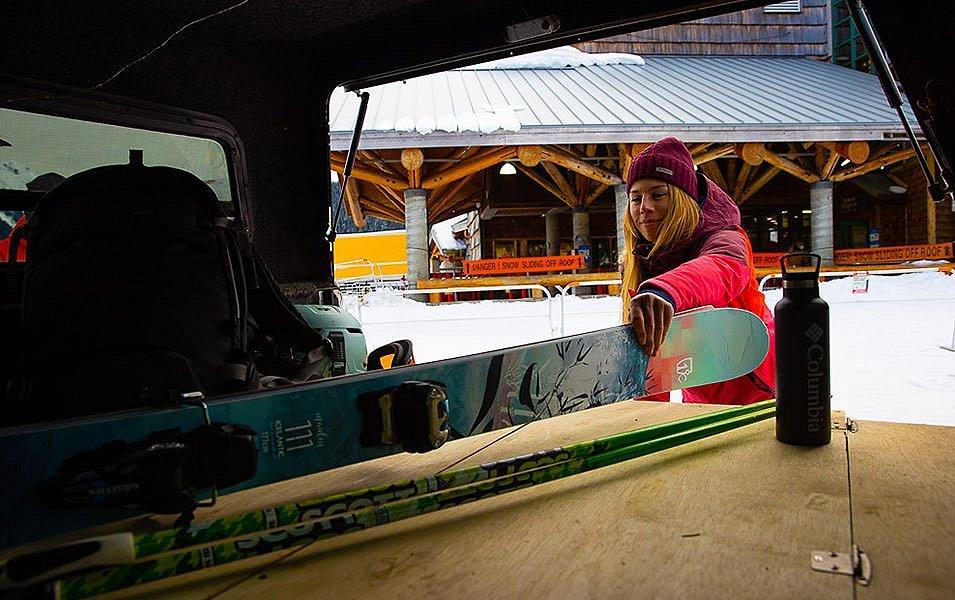 A woman in a reddish pin jacket pulls a set of skis out of her car with a ski lodge in the background. 