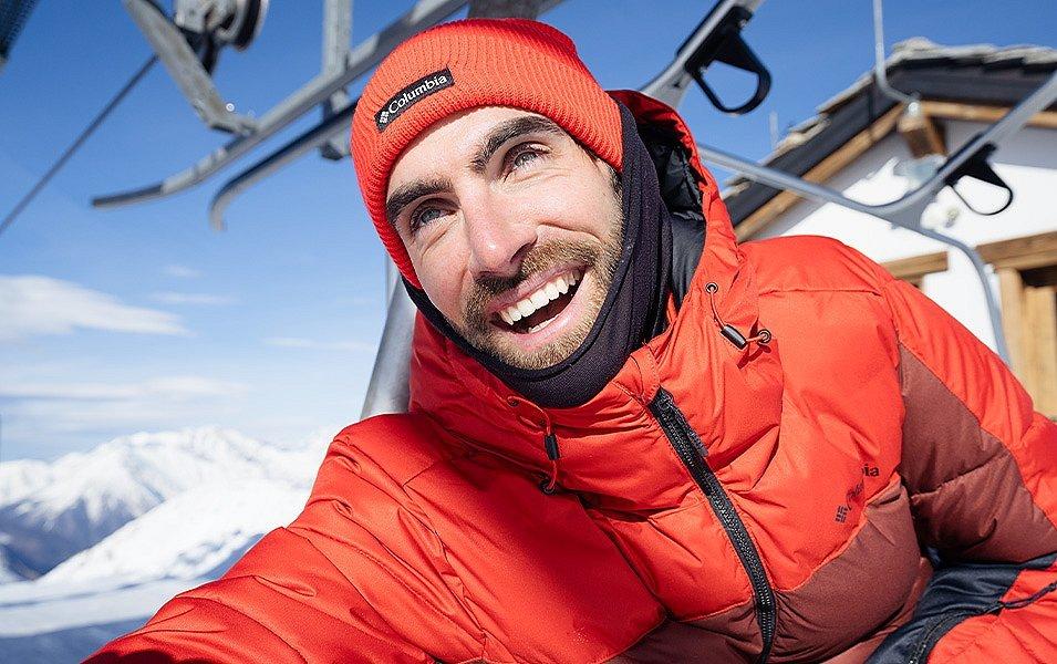 A man in a red Insulated Columbia Jacket sits on a chairlift on a snowy mountain. 